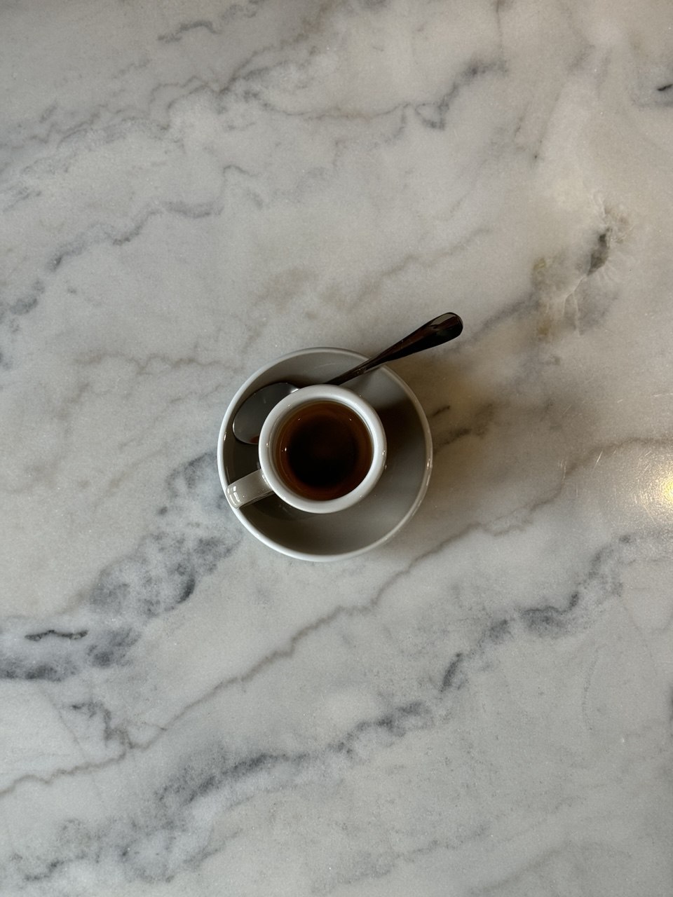 A single espresso in a white cup with a spoon rests on a matching saucer atop a marbled, white and gray table.