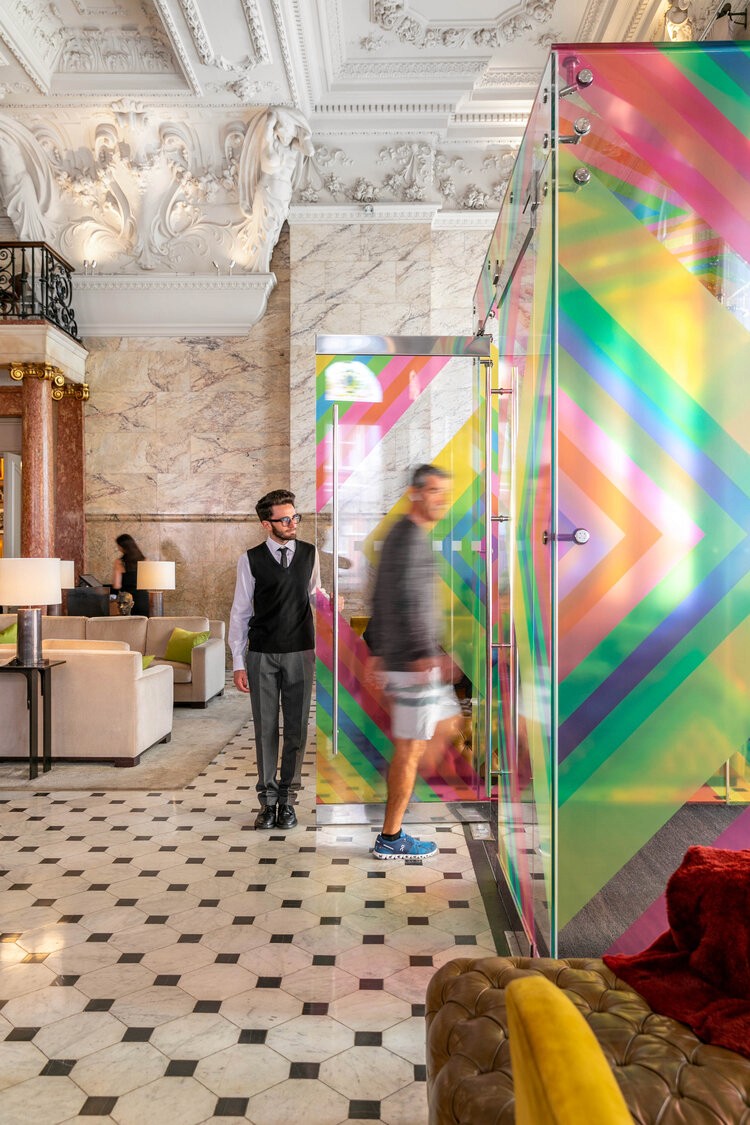 A hotel lobby with marble flooring, intricate ceiling details, and a colorful translucent panel near the seating area.