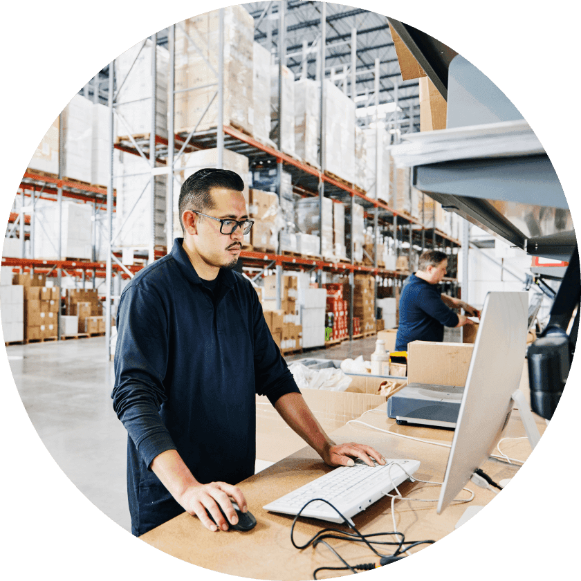 A man with glasses working at a computer in a warehouse with shelves stocked with boxes in the background. Another worker is visible in the background, organizing items.
