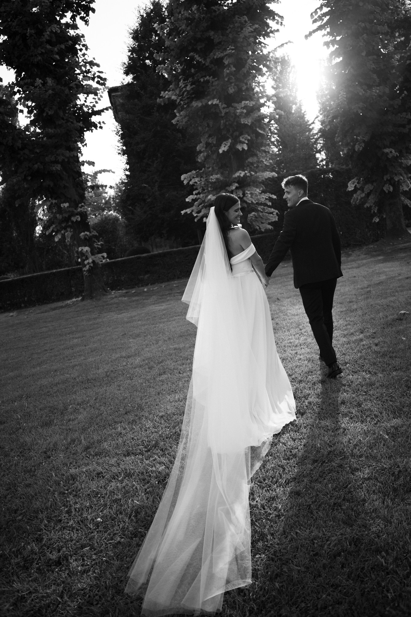 Sposi camminano nel giardino di Villa Negri, il velo della sposa si allunga dietro di lei tra gli alberi in controluce