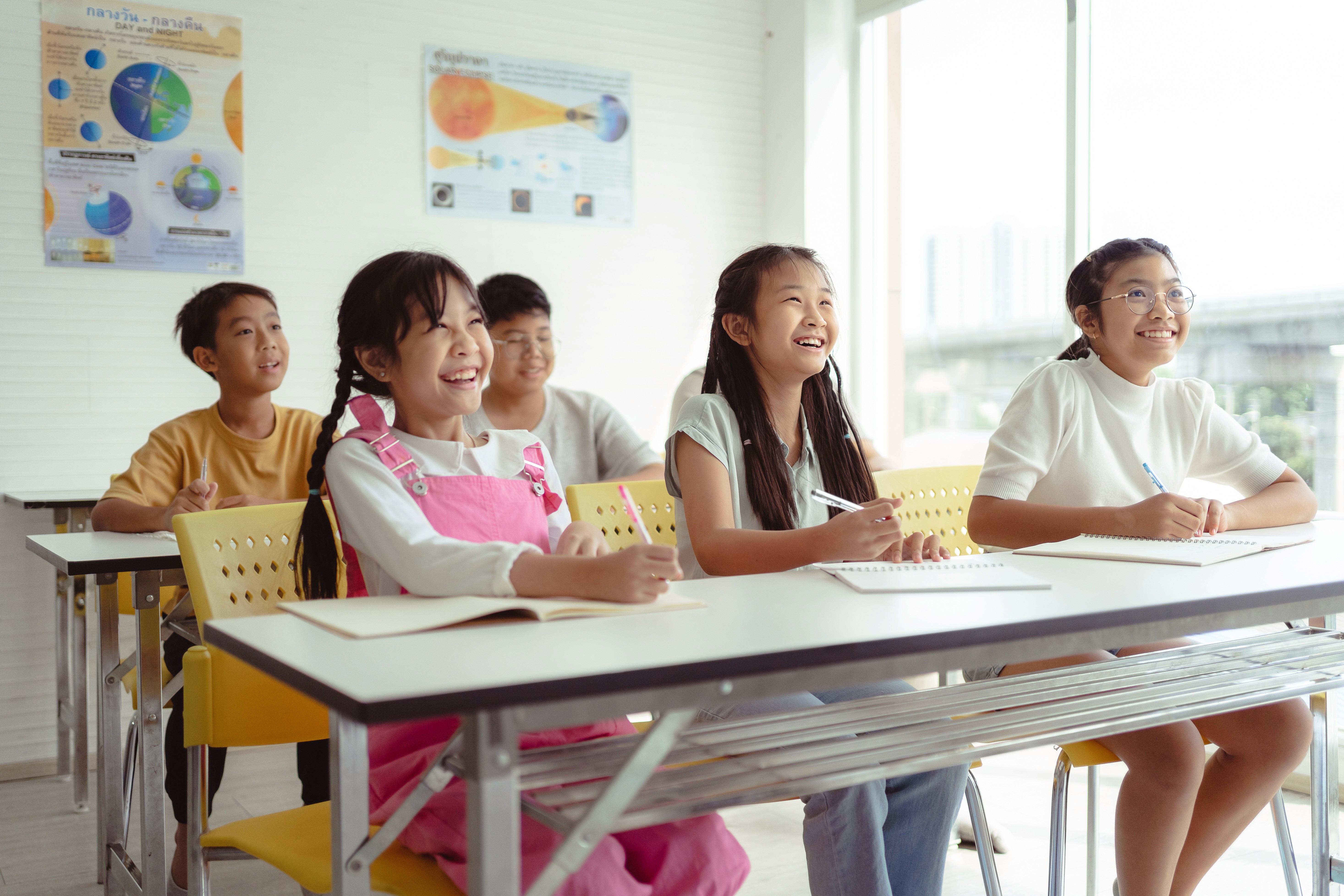 A diverse group of children are seated at desks in a bright classroom, smiling and taking notes, with educational posters visible on the wall.