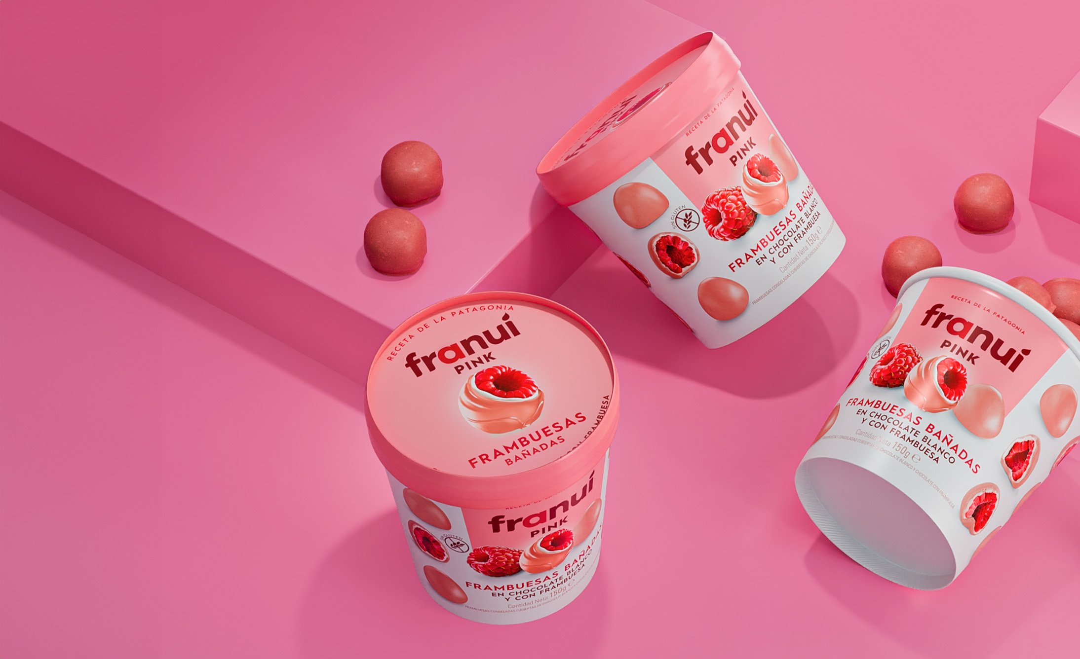 Three tubs of ice cream labeled "Franuí Pink" with raspberry images and pink packaging are on a pink surface. Several chocolate-covered raspberries are scattered.