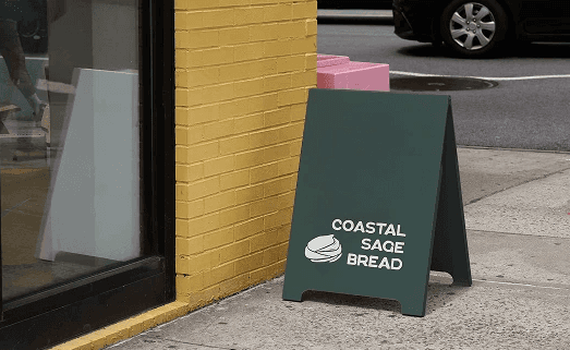 deep kelp green sandwich board with the coastal sage bread logo on the front. It's sitting in front of a yellow building 