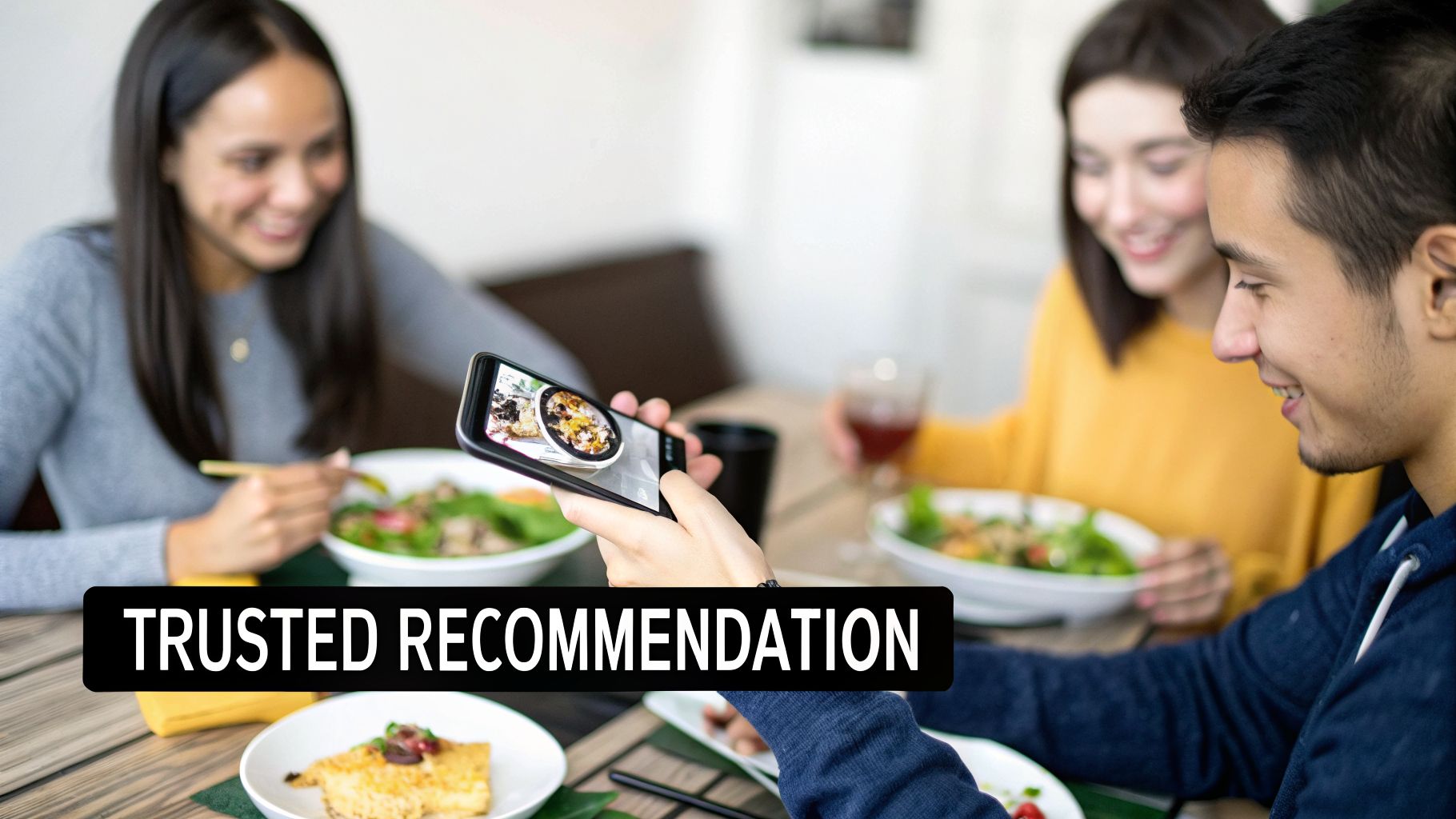 Three friends at a restaurant, one person takes a picture of food with a smartphone, smiling.