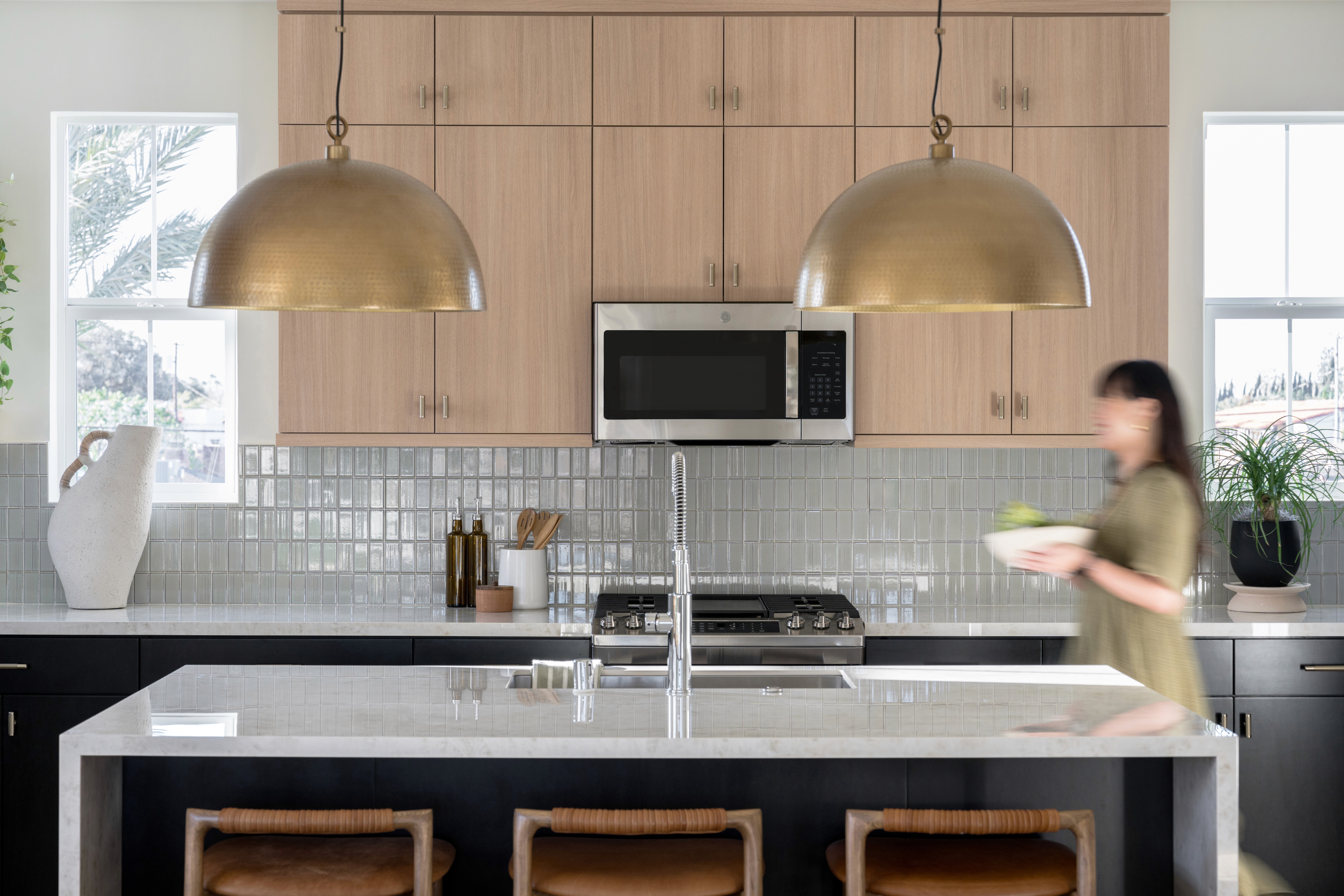 Kitchen island with large, brass pendant lighting, woven counter stools, light wood upper cabinets, sea green tile backsplash, and black lower cabinets