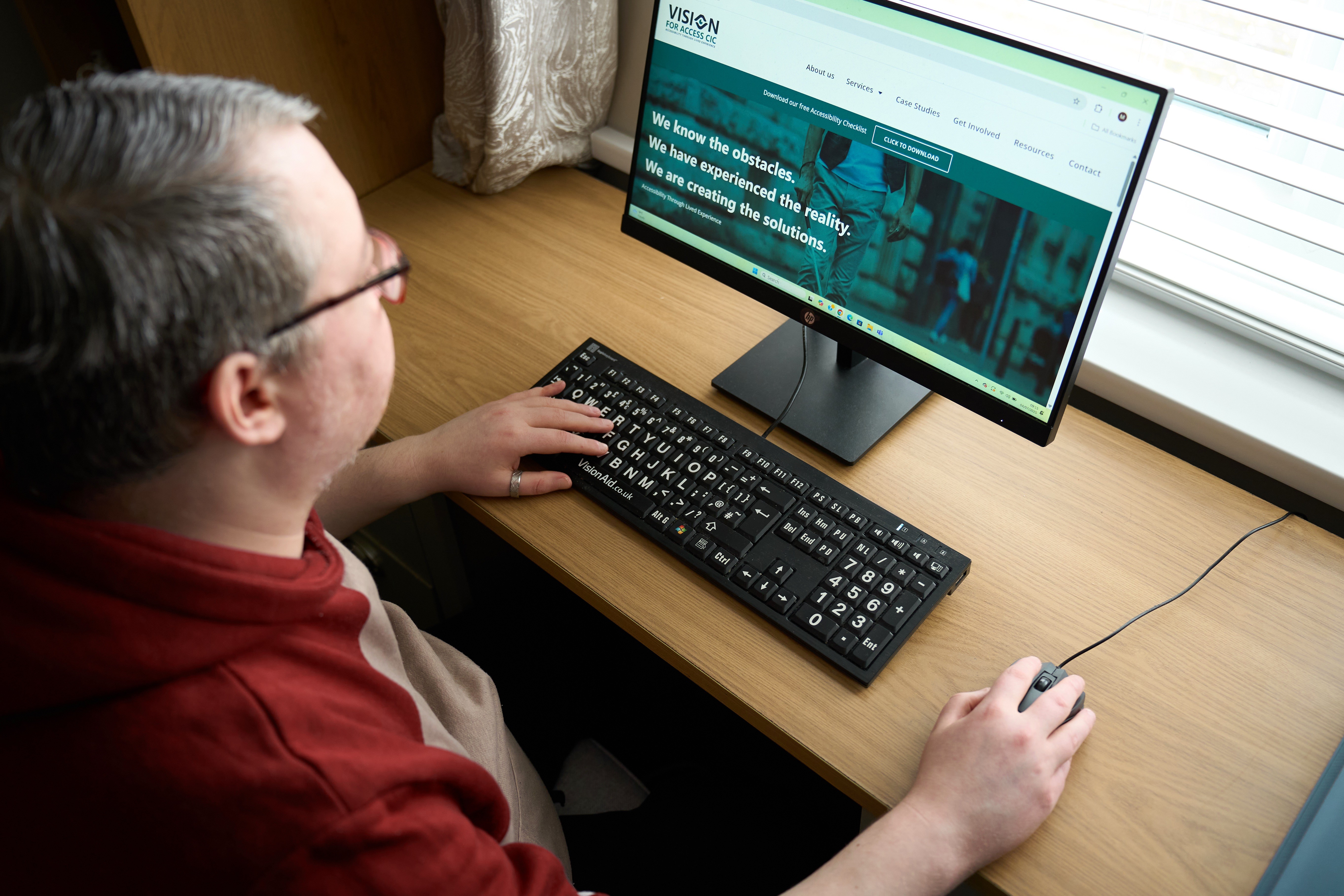 An image of Mark, sat at a desk on a computer. His left hand is on the keyboard, his right on the mouse. He is looking at a website.