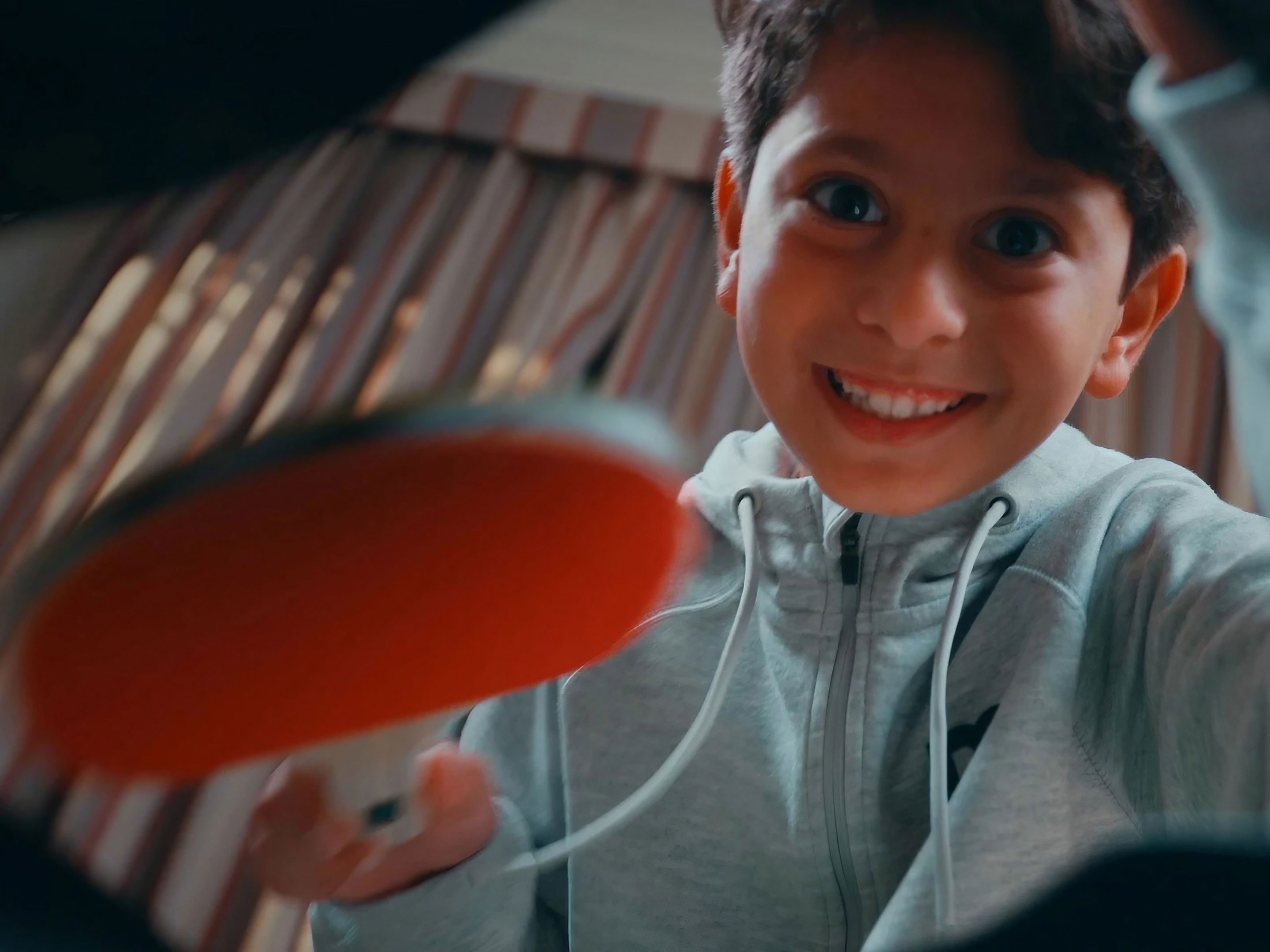 A smiling boy in a grey hoodie tucking a red table-tennis paddle into his school bag, from the 2023 campaign.