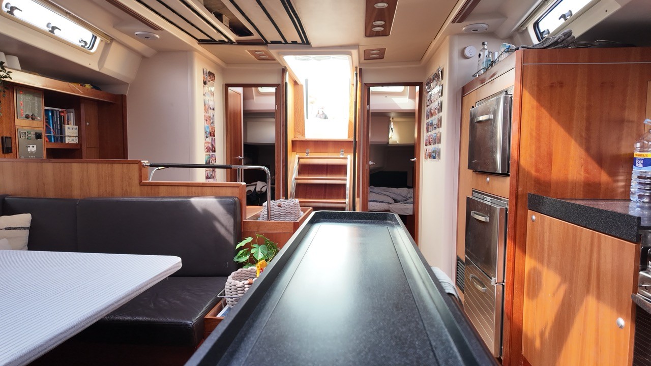 Beneteau Clipper 361 main saloon interior showing the stainless steel galley sink with chrome tap, white worktop, mahogany cabinetry throughout, nav station with red panel, analogue gauges and chartplotter, teak-and-holly cabin sole, blue fabric U-shaped dinette seating with folding table, and a flat-screen TV mounted in the forward bulkhead