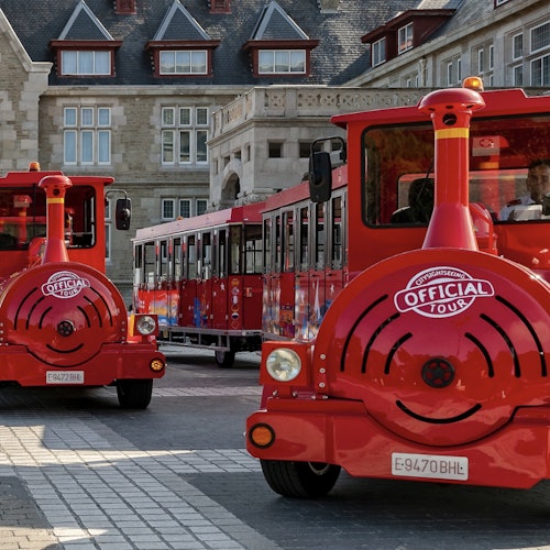 Two red tour trains with "Official Tour" logos parked outside a large, ornate stone building with multiple windows and gables.