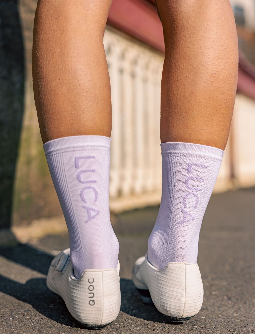 Close-up-rear-view-of-a-person's- lower-legs-wearing-light-lavender- cycling-socks-that-have-the-word- LUCA-knitted-vertically-on-the-back-of-the-calf-The-person-is-also- wearing-white-cycling-shoes-with- the-QUOC-brand-visible-on-the-heel