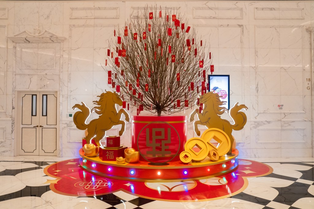 A festive display featuring a decorated tree with red envelopes hanging from branches, flanked by two golden horse sculptures and surrounded by gold ingots, set against a marble wall.