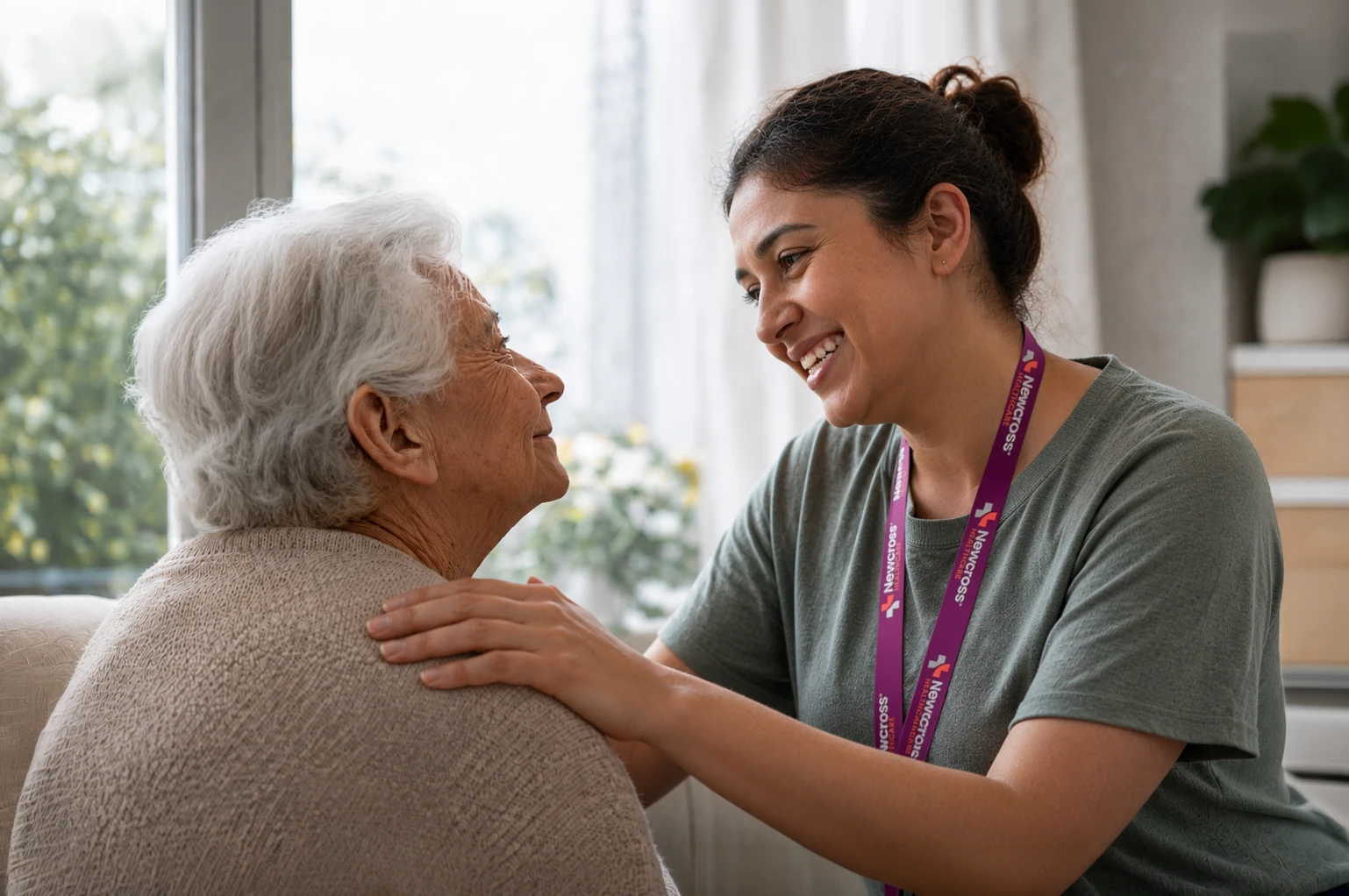 A Newcross care worker in casual clothing smiles warmly at an elderly woman in her home, resting a hand gently on her shoulder as sunlight comes through the window.