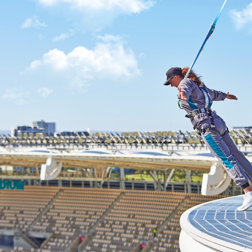 Person in safety gear leans off a rooftop, attached to a harness, with an empty stadium and clear sky in the background.