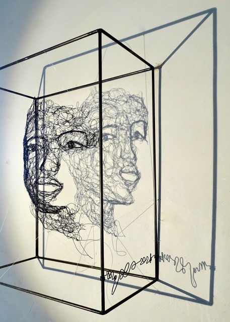 Sculpture en fil de fer représentant le visage d'une femme dans un cadre en fil de fer, vue de profil avec son ombre projetée sur le mur