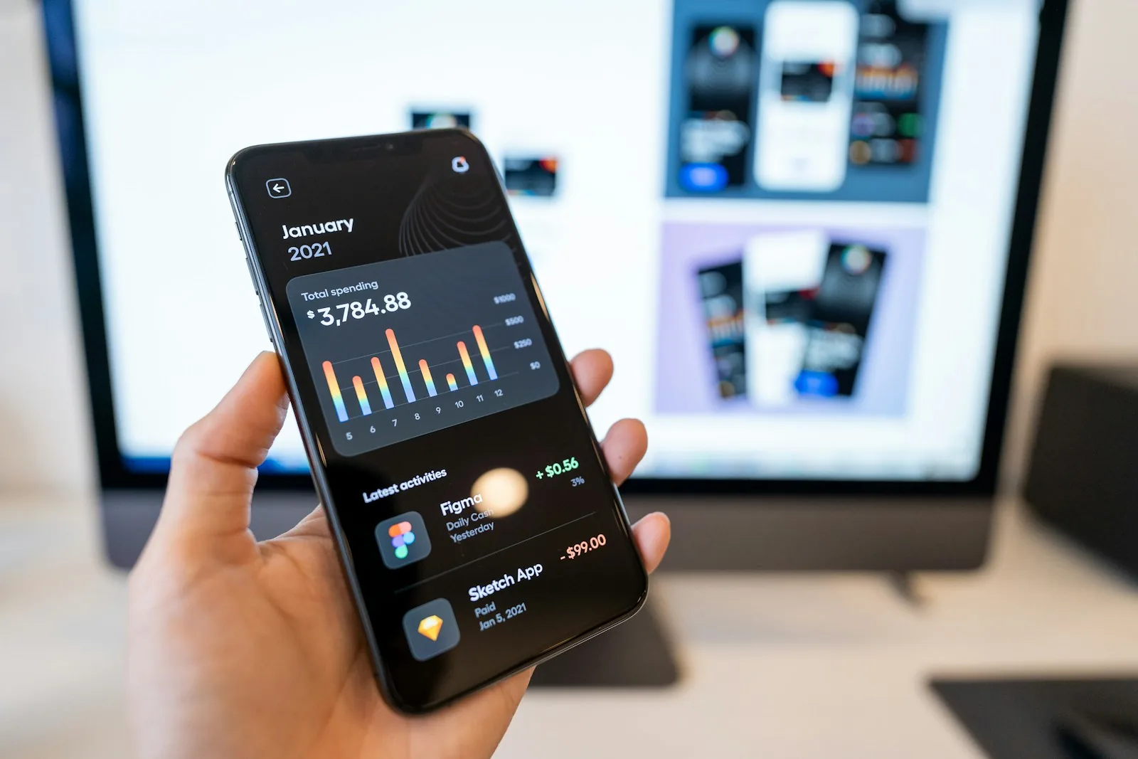 Hand holding a smartphone showing a dark-mode spending app with a monthly bar chart and recent transactions, in front of a monitor displaying mobile app screen mockups
