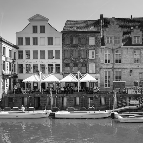Black-and-white photo of a canal with boats in front of a row of old buildings and a restaurant with outdoor seating.