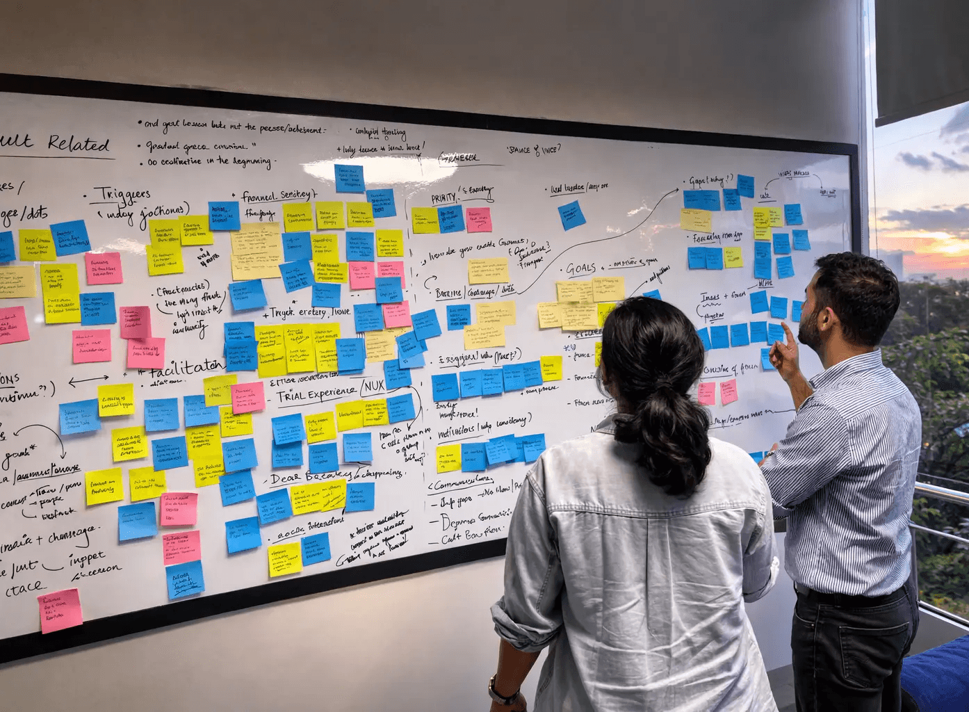 A man in a brown blazer and a woman in a light sweater sit at a desk with a laptop, discussing something. The room is dimly lit, creating a focused atmosphere.