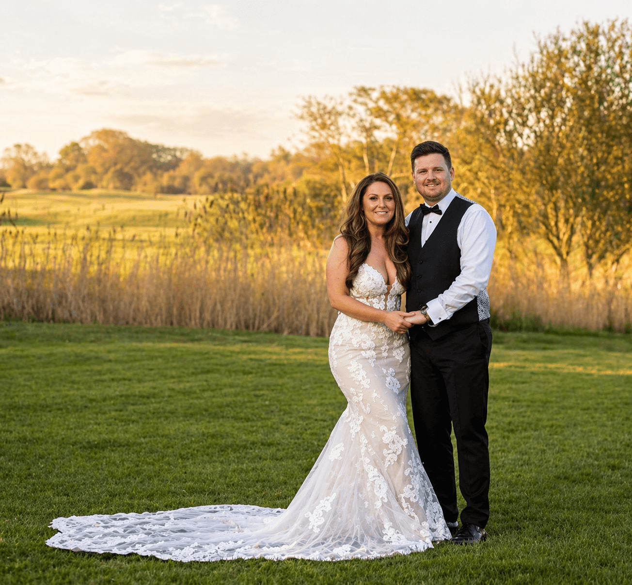 A smiling bride and groom stand holding hands on a green lawn with trees and tall grass in the background at