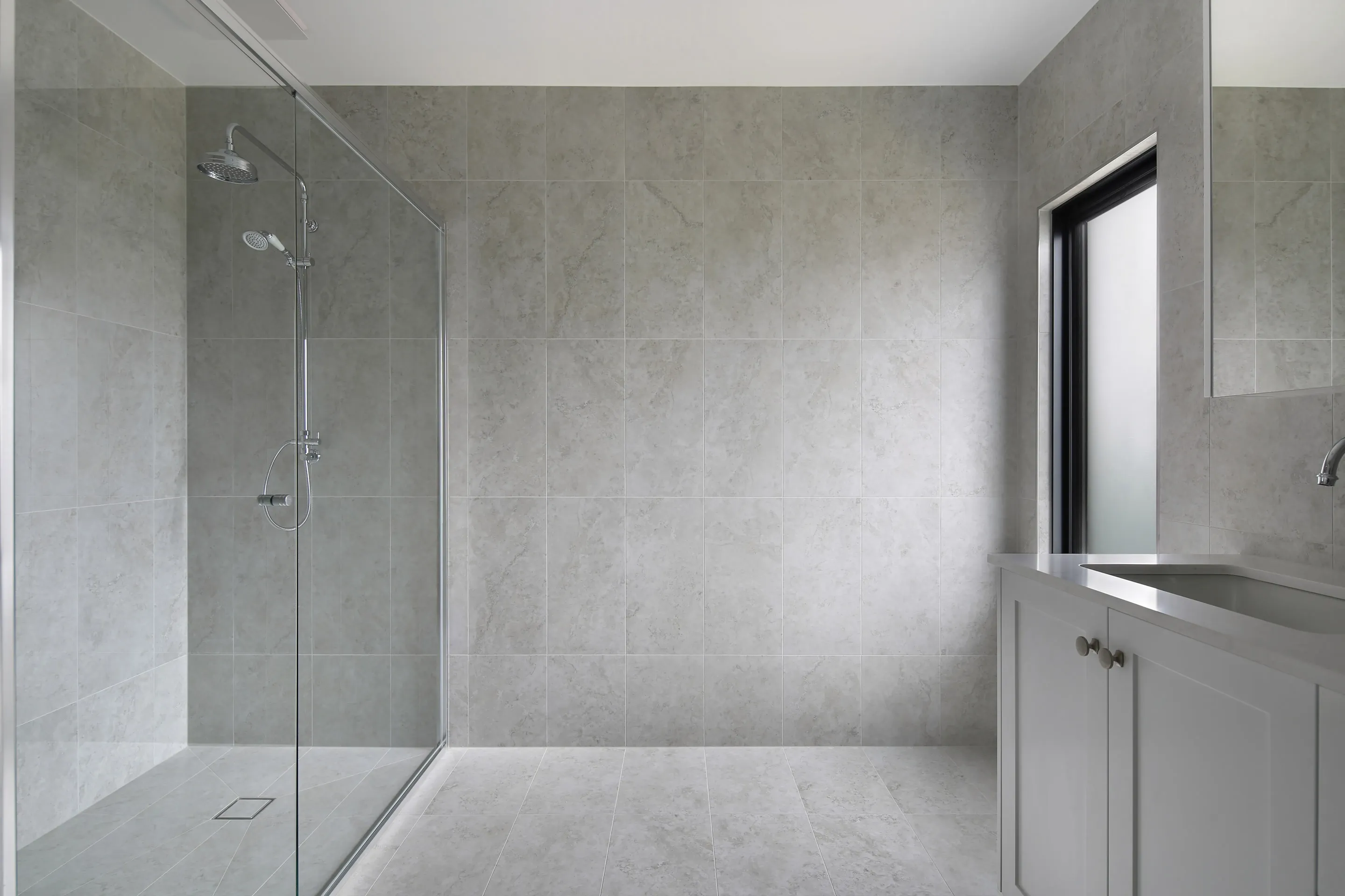 Bathroom with grey stone-look tiles, a walk-in glass shower with a chrome rail shower, a white shaker vanity and a frosted black-framed window