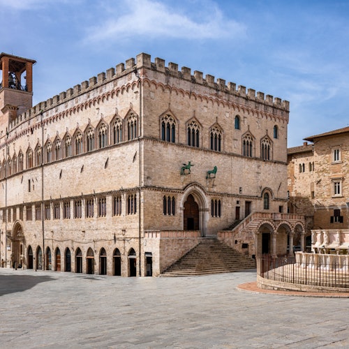 Historic stone building with arched windows and stairs, adjacent to a fountain in a clear open square under a blue sky.
