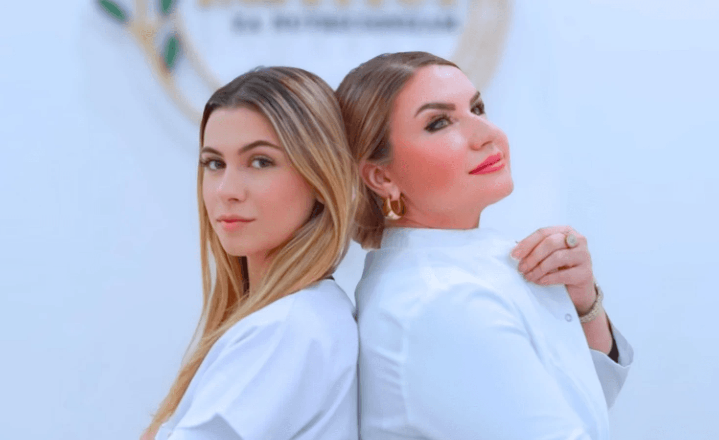 A young woman with curly hair and glasses smiles warmly while sitting in a sunlit room filled with various potted plants, creating a cozy and inviting atmosphere.