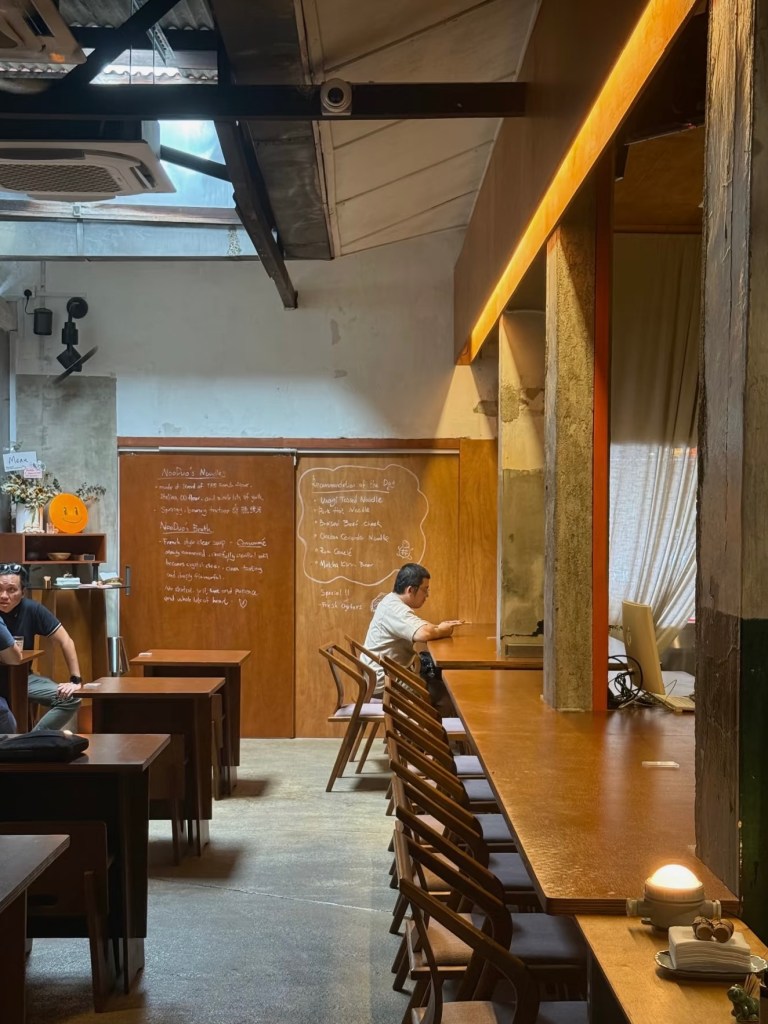 Interior of a cafe featuring wooden tables and chairs, with a person sitting at a table using a device. A chalkboard with handwritten notes is visible on the wall.