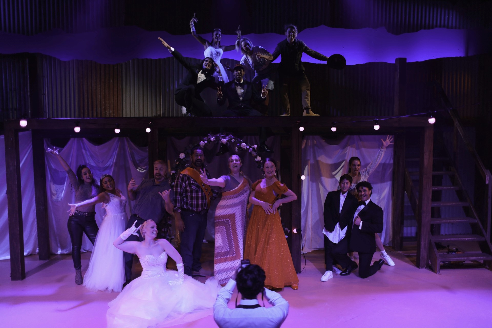An ensemble of performers pose joyfully for a staged group photo during a celebratory wedding scene. Some are positioned on an upper platform while others kneel or stand below, dressed in gowns, suits, and vibrant costumes. A photographer faces the group in the foreground as purple lighting bathes the stage.