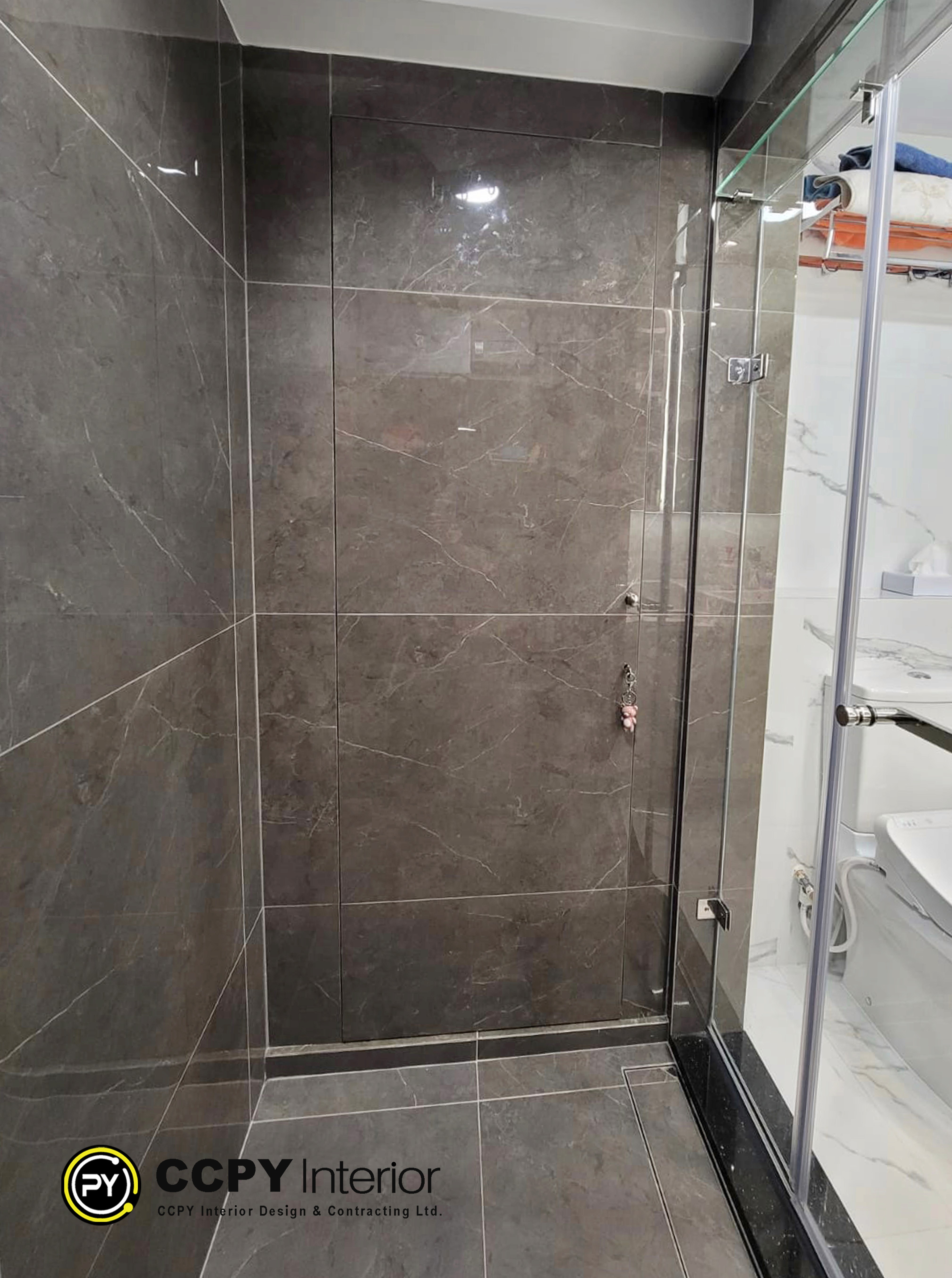 The selected region showcases a modern shower enclosure featuring dark stone-texture porcelain wall tiles, a glass panel partition, and a sleek chrome shower fixture setup.Design ElementsWall Tiles: Large-format, dark gray porcelain tiles with realistic white marble veining.Enclosure: Minimalist, frameless tempered glass partition wall that maximizes light flow.Fixtures: Polished chrome shower mixer valve paired with a modern hand-held shower wand.Corresponding SpecificationsAccording to the accompanying design notes, this bathroom features:Wall & Floor Tiles: Dark stone-texture porcelain tiles.Fixtures: Kusatsu bathroom heater and ventilation fan, paired with a TOTO smart toilet.