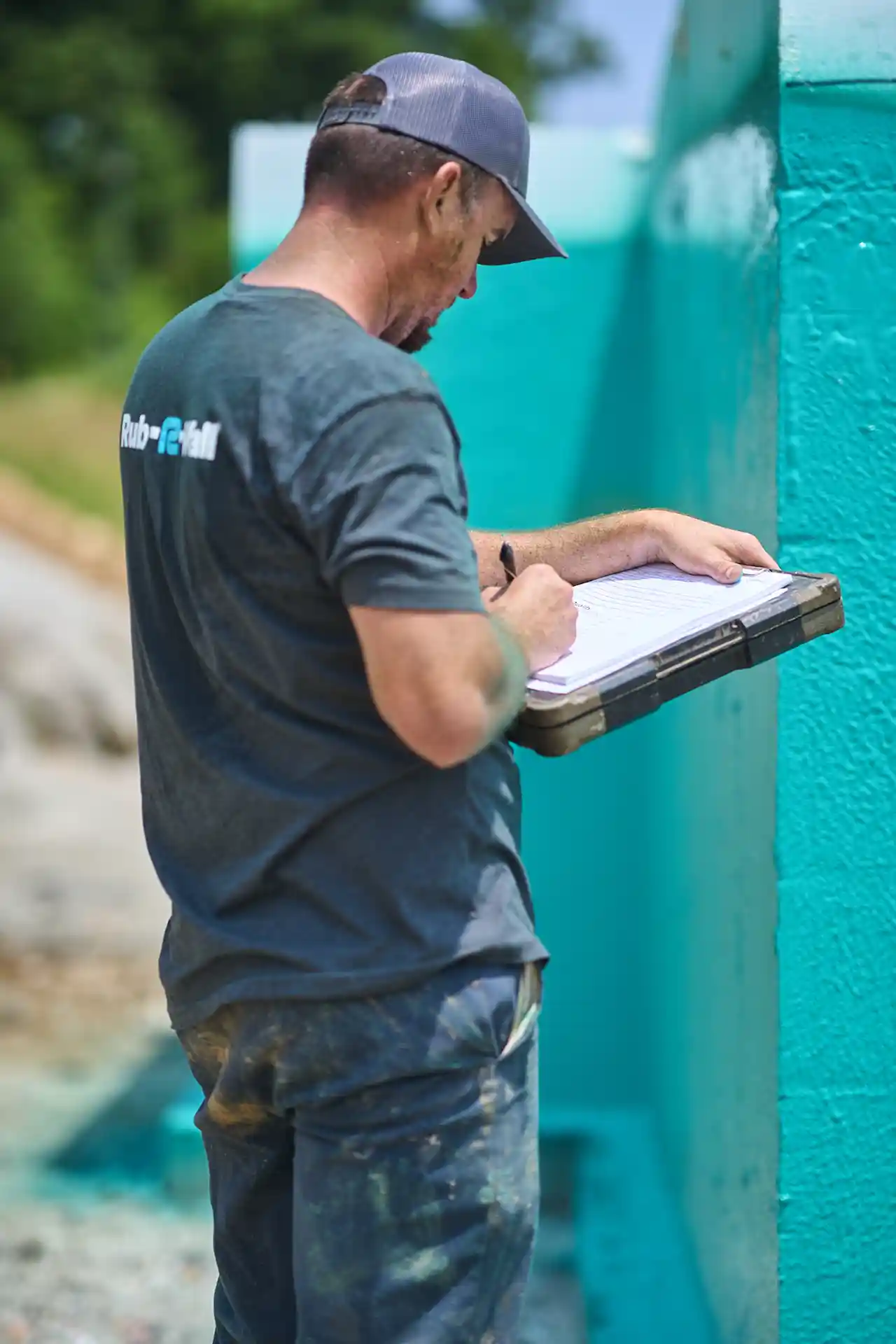 A man in a Rub-R-Wall t-shirt taking job notes on a clipboard after he finished waterproofing a house