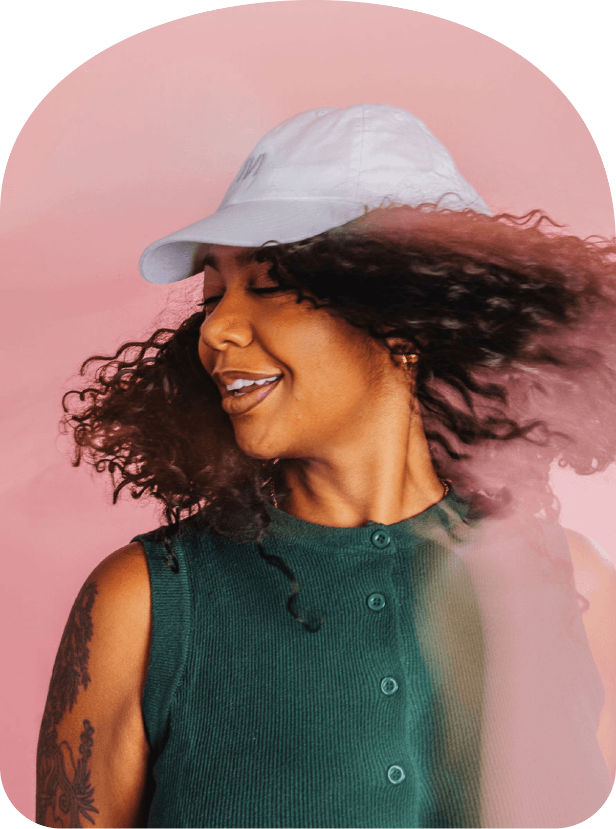 Woman with curly hair wearing a white cap, turning her head mid-motion against a soft pink background.