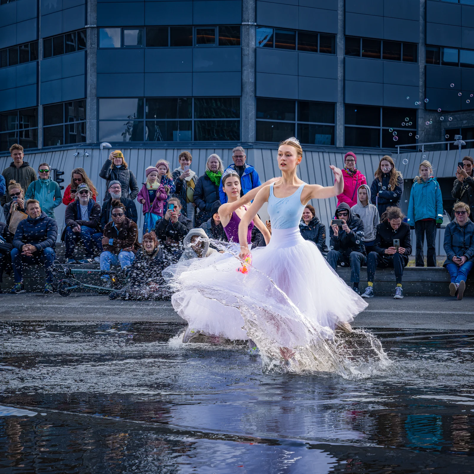 Two ballerinas in tutus perform a dynamic dance routine, splashing through a shallow pool of water on an outdoor public square during Reykjavík Culture Night. A diverse crowd of spectators sits and stands in the background watching the live performance in front of a modern city building.