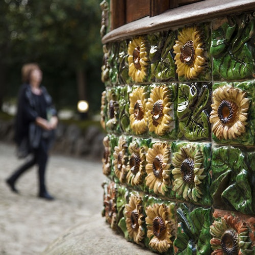 Una mujer camina junto a una pared decorada con coloridos azulejos de cerámica que presentan motivos de girasoles en relieve.