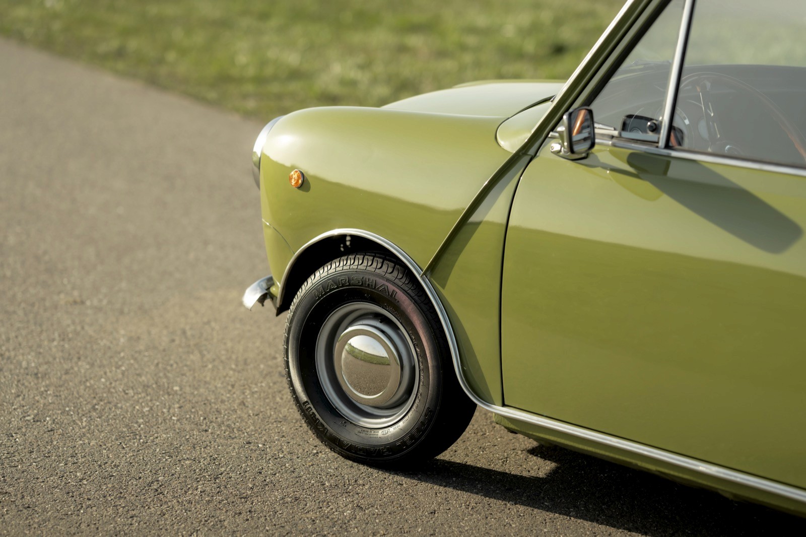 Close-up of a green vintage car's front wheel and fender on a gravel road, with grass in the background.