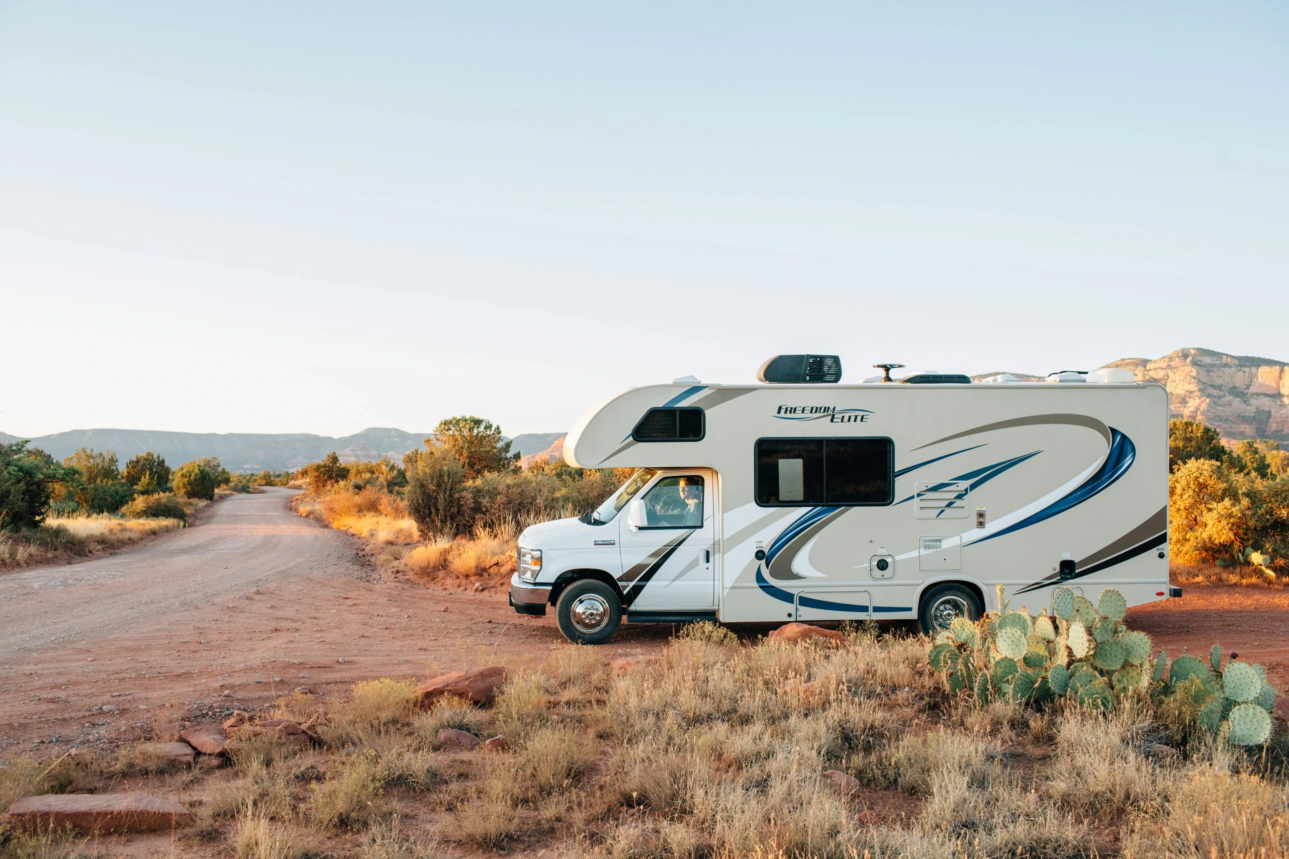 Blurred white Class C motorhome parked in a desert landscape at sunset with cacti and mountains