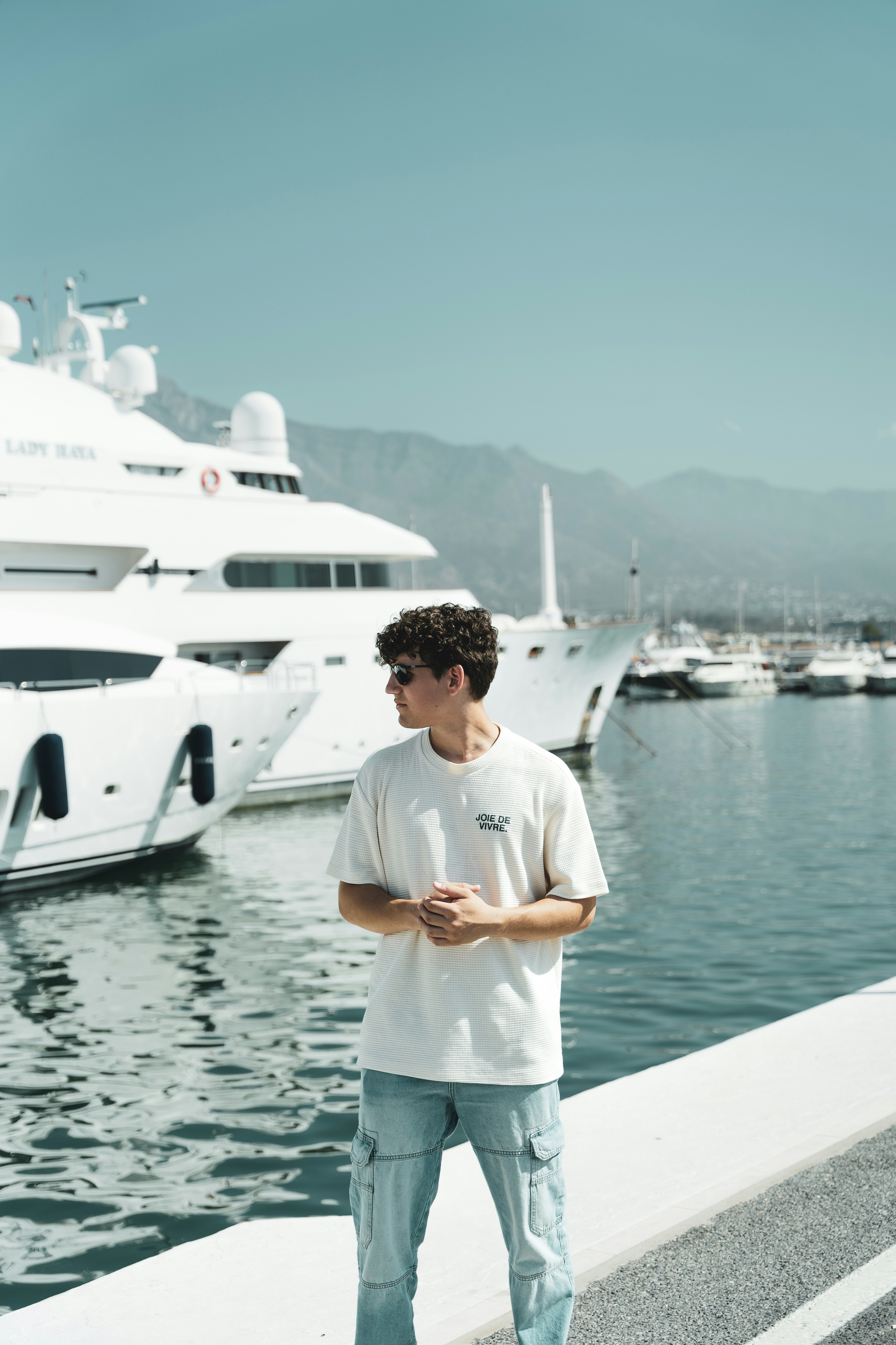 An outdoor photograph of a young man with curly dark hair wearing sunglasses and a white t-shirt. He stands with his hands clasped in front of him, looking toward the left side of the frame. The background features a large white yacht and other boats docked in a harbor with coastal mountains visible in the distance
