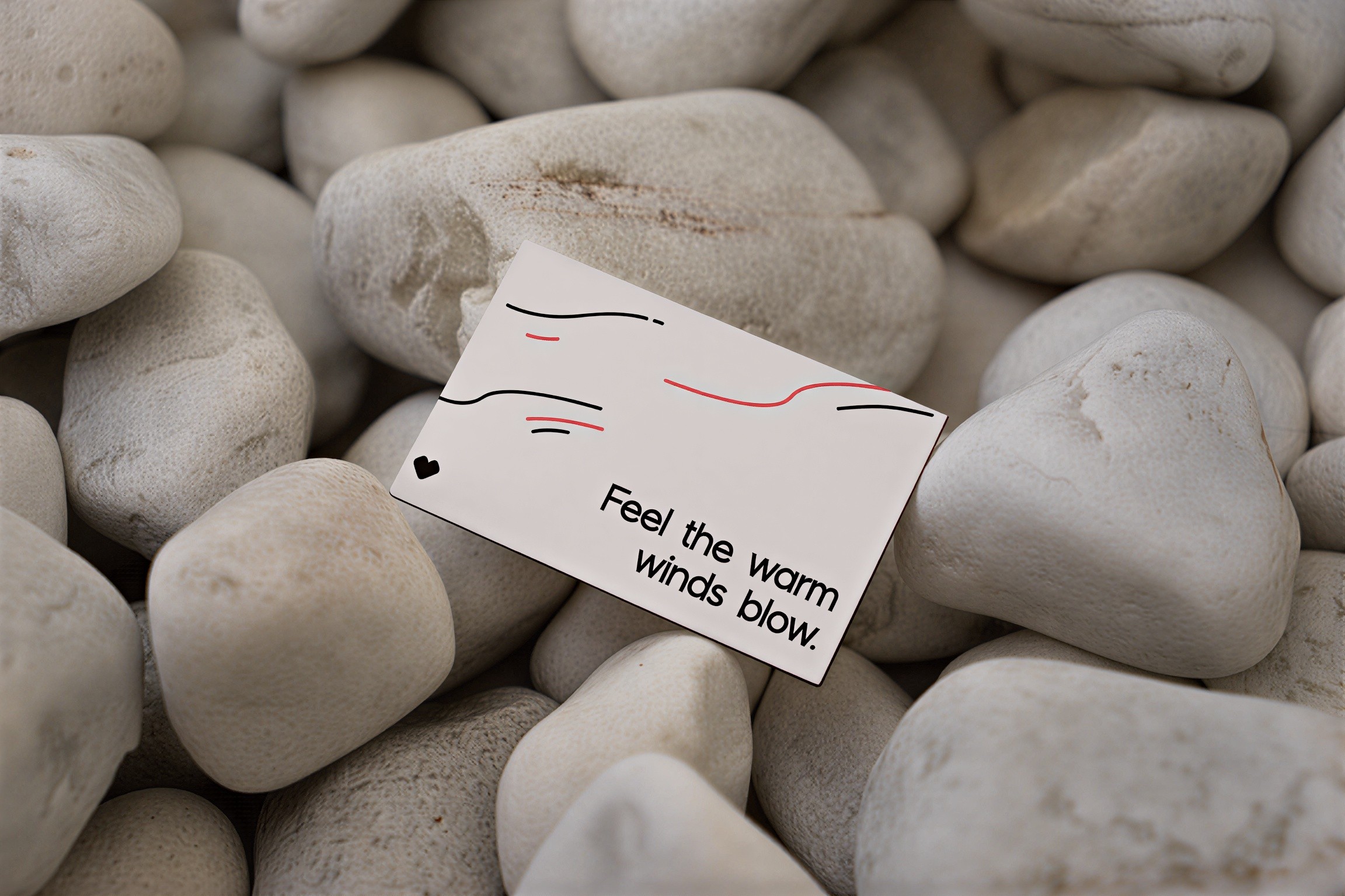 A white card with abstract line art and the text "Feel the warm winds blow" rests among a pile of smooth, white pebbles.