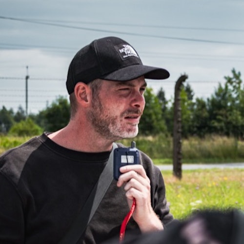 Man die buiten een portofoon vasthoudt, draagt een zwarte pet en shirt met groen en een hek op de achtergrond.