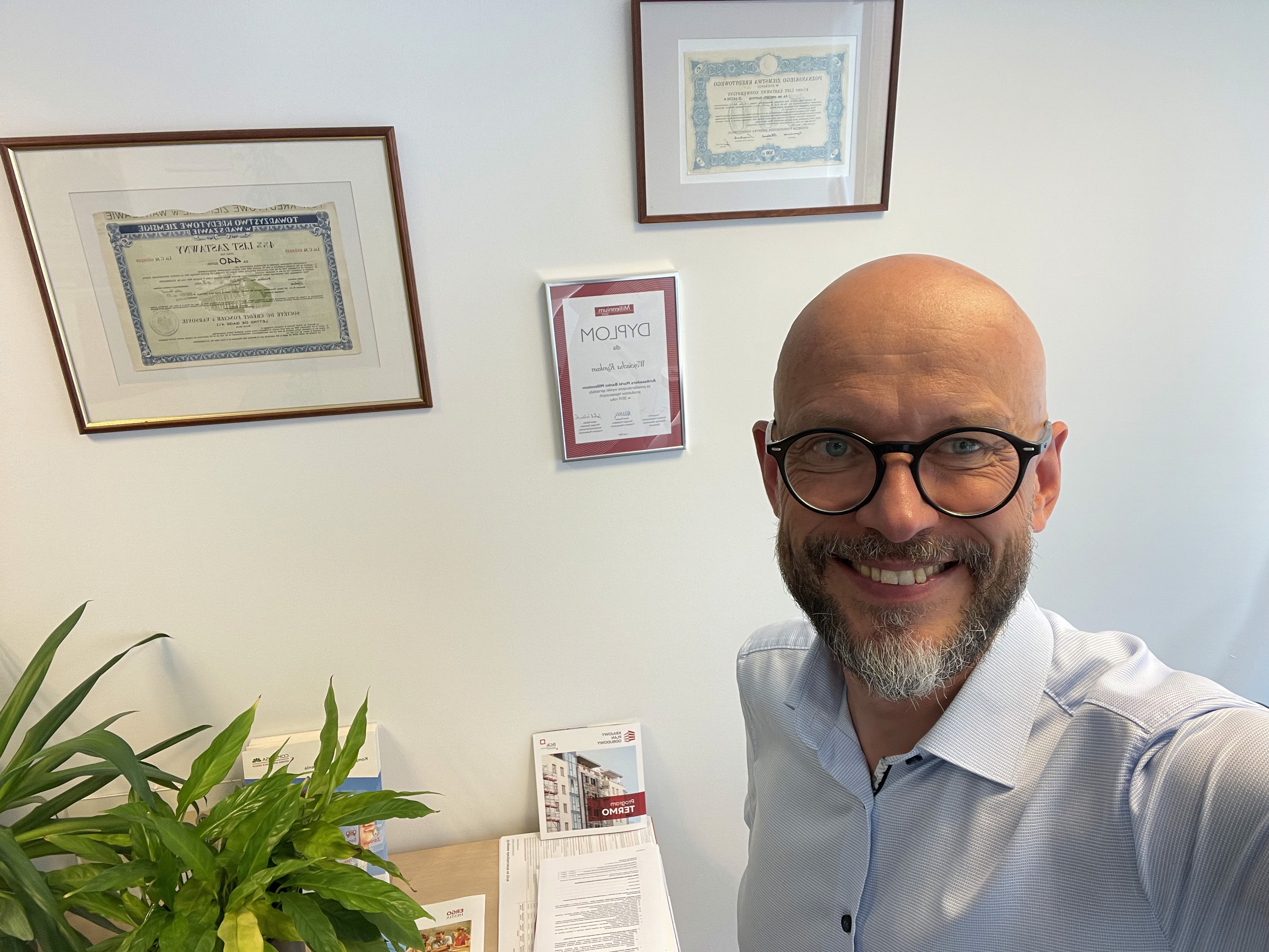 A stylish, modern lounge area featuring pastel green sofas, tan chairs, round wooden tables, and ceiling plants, with large windows and sheer curtains.