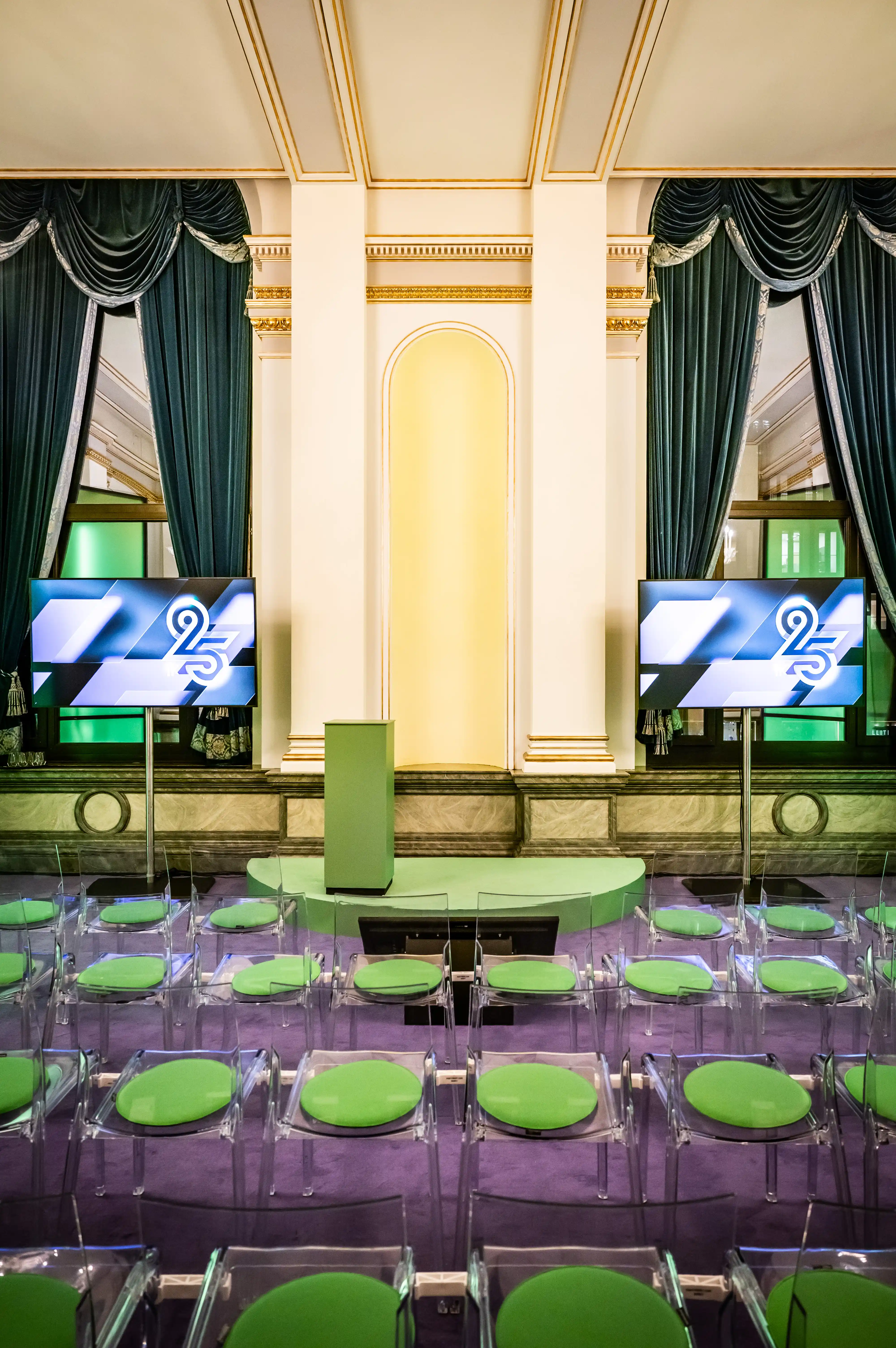 Ballroom with transparent chairs with green cushions and 25th anniversary screens between drapes.
