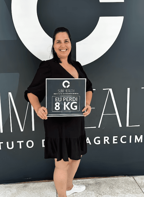 A smiling woman with her arms crossed, standing against a dark green background. She has long, dark hair.