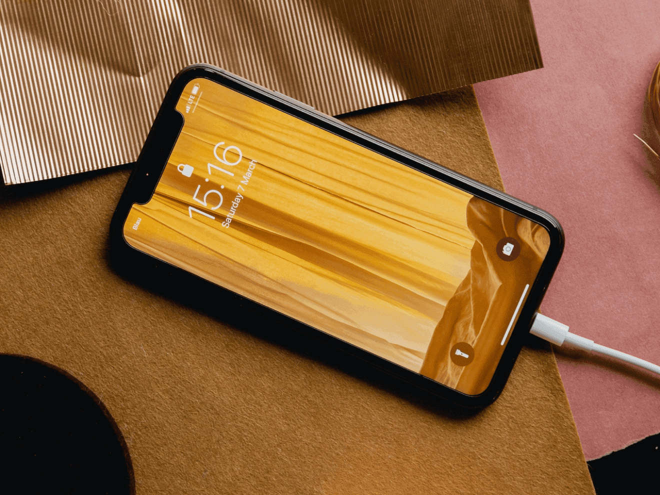 Smartphone with a yellow fabric wallpaper displayed on the lock screen, placed on a textured brown and pink surface with metallic accents, connected to a charging cable.