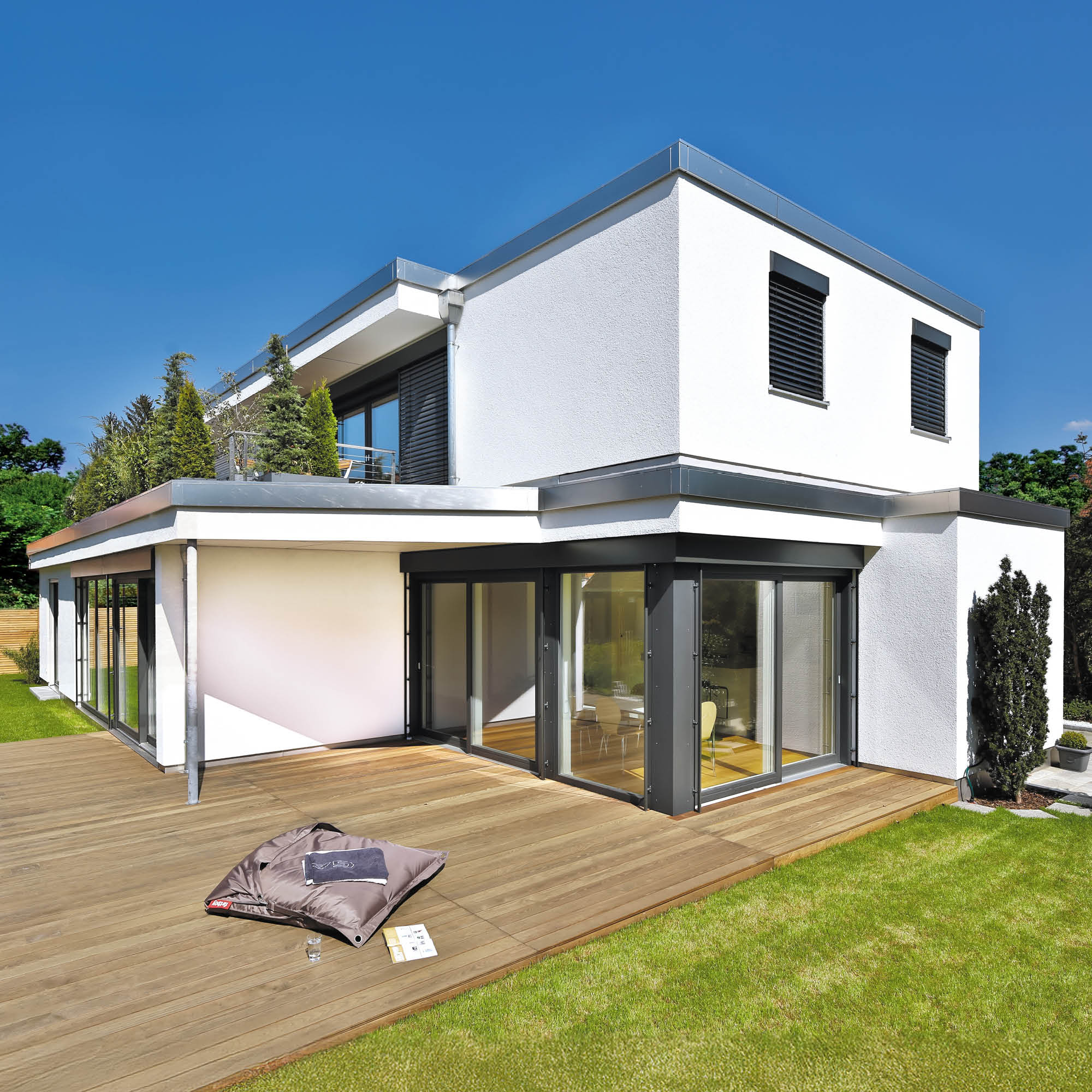 Modernes weißes Haus mit großer Holzterrasse, Glasfronten, begrüntem Dach und viel Grün im Garten.