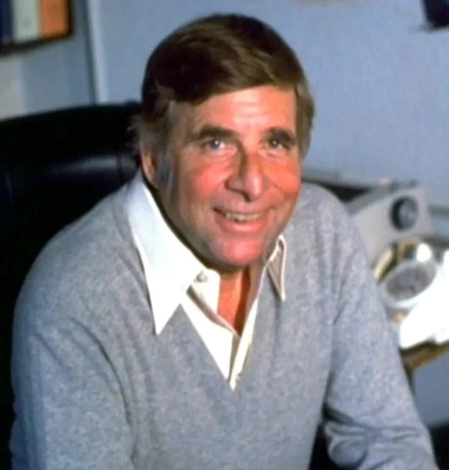 An elderly man with short, dark hair and a friendly expression is seated indoors wearing a light gray sweater over a white collared shirt, with a glimpse of an old-fashioned rotary telephone in the background.