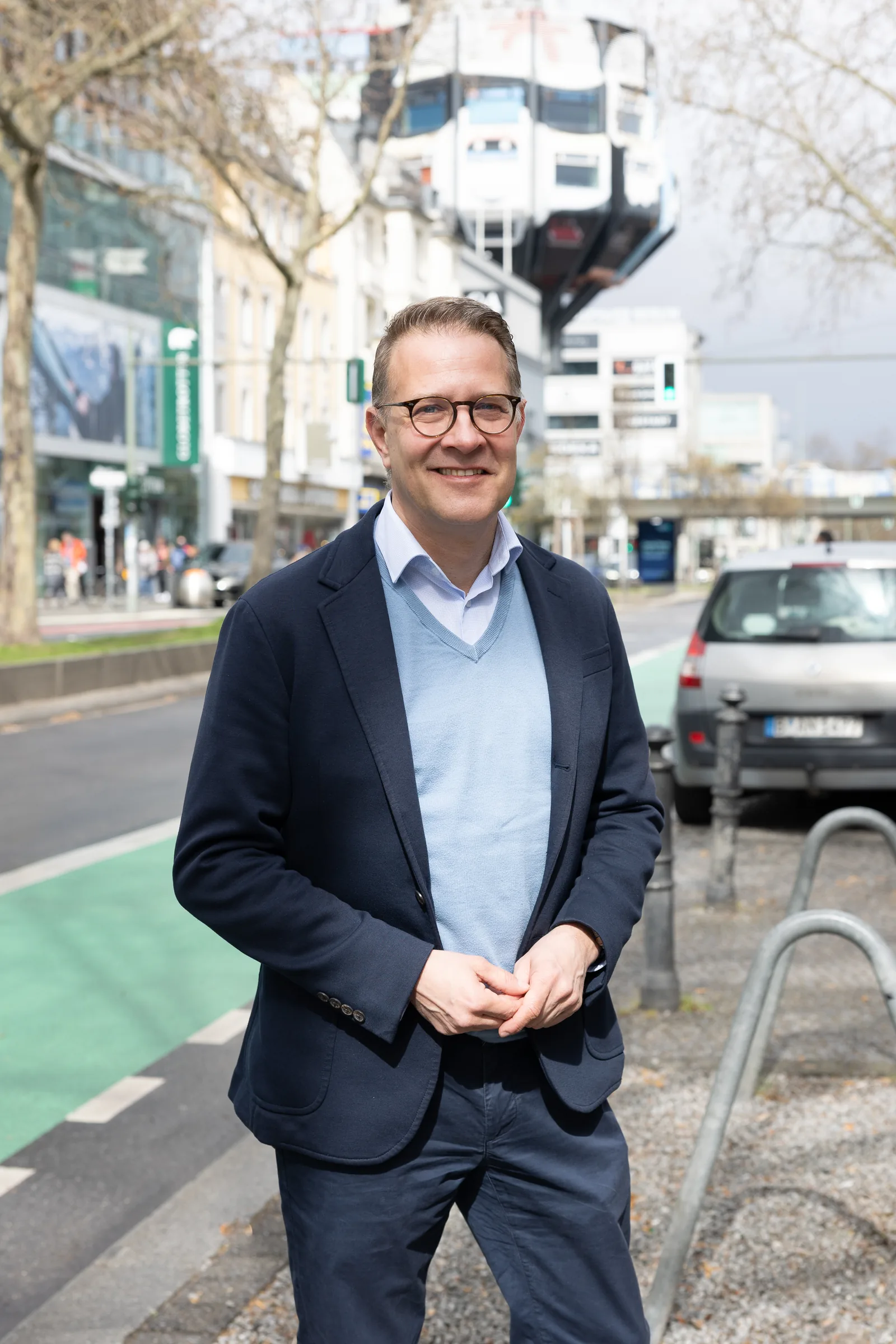 Adrian Grasse steht lächelnd auf einer Straße in seinem Heimatbezirk Steglitz-Zehlendorf, im Hintergrund ist belebte, urbane Architektur zu sehen.