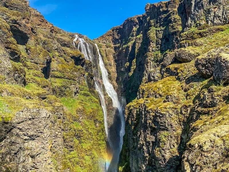 Tall waterfall at Glymur in Iceland flowing through steep canyon walls made of dark, thin basalt layers, with visible stacked lava formations and green vegetation along the cliffs.