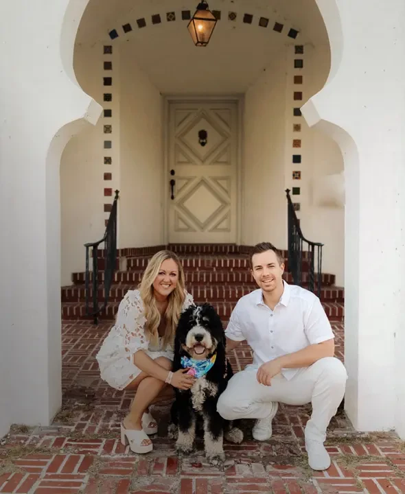 Founders Matt and Christen Hildesheim with their dog Luna sitting on brick steps outside a home with an arched entryway.