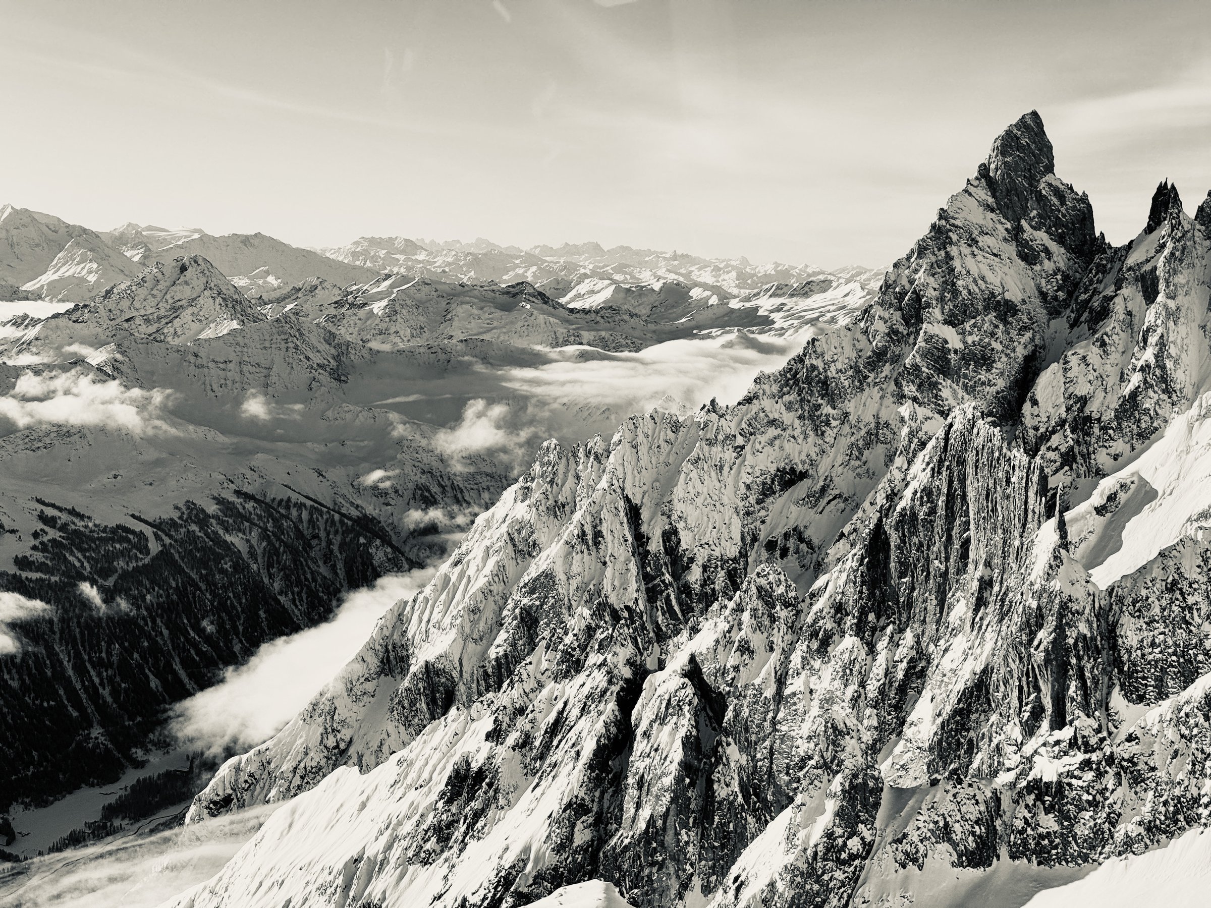 Black-and-white view of jagged snow-covered peaks and a cloud-filled valley from the Mont Blanc massif above Courmayeur, Italy