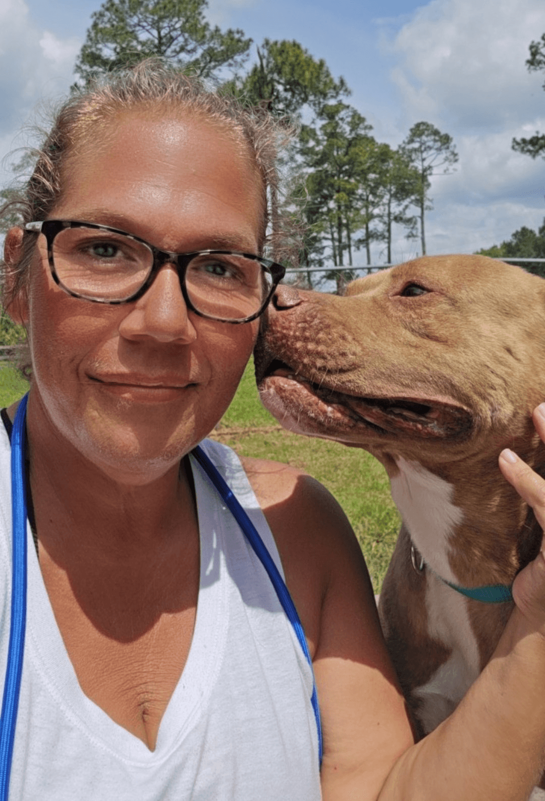 A person with glasses smiles at the camera while a dog leans in for a kiss in a sunny outdoor setting.