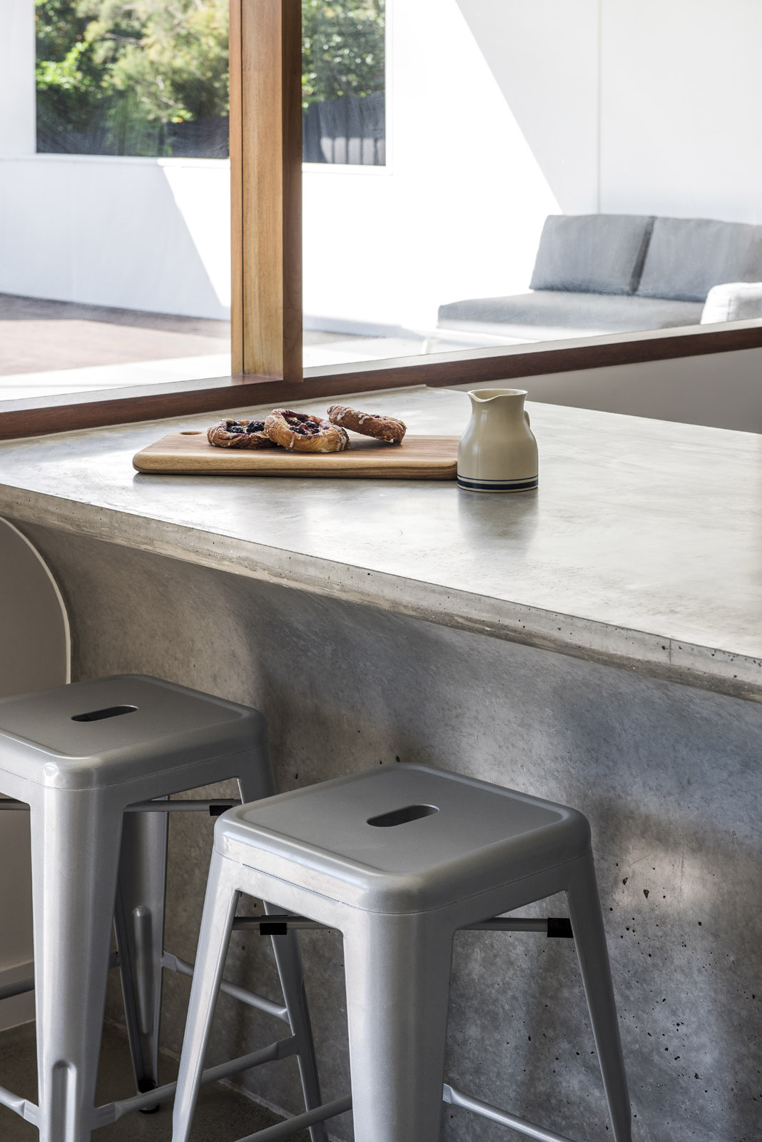 Close-up of concrete kitchen bench with pastries, ceramic jug, and bar stools near window