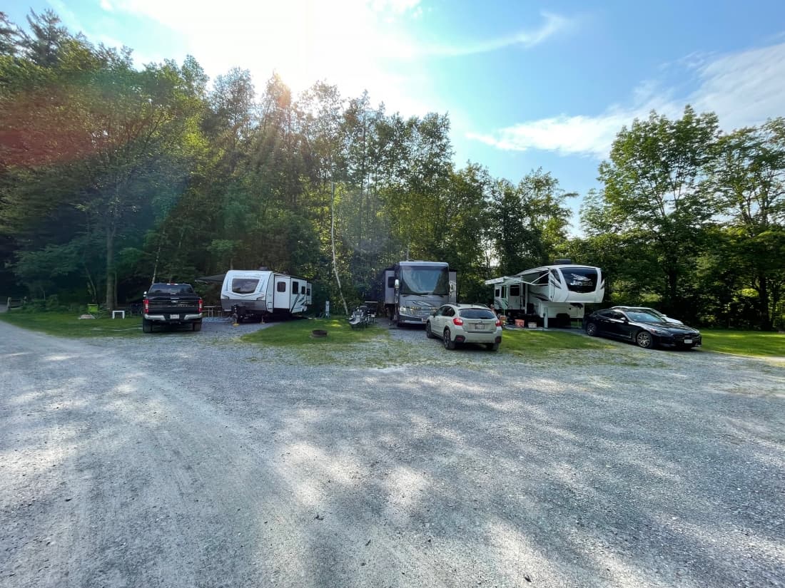 Row of back-in RV sites with full hook-ups, gravel pads, and lush green forest backdrop